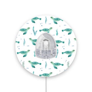 Può includere: Un diffusore rotondo bianco con un motivo di tartarughe marine verde acqua e verde. Il diffusore ha un cavo bianco e un coperchio in plastica trasparente.