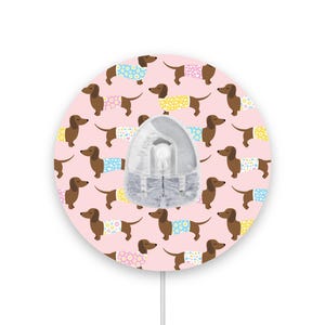 Peut inclure: Un autocollant mural rond rose avec un cordon blanc. L'autocollant présente un motif répétitif de teckels bruns portant des vêtements colorés avec des motifs floraux.