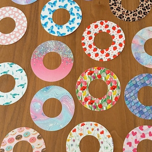 May include: A set of 14 round, colorful, patterned stickers with a hole in the center. The stickers feature various designs, including butterflies, hearts, donuts, flowers, glitter, and animal prints.