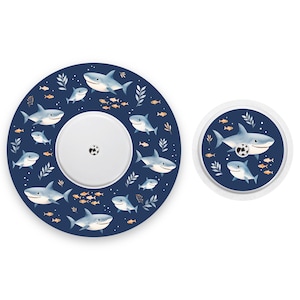 Può includere: Un set di due coperture rotonde blu e bianche per dispositivi medici con un motivo a squalo e pesci. La copertura più grande ha un centro bianco con un piccolo foro al centro. La copertura più piccola ha un bordo bianco e un centro blu.