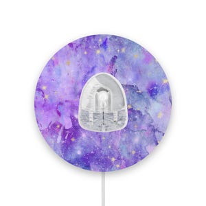 Peut inclure: Un dispositif médical avec un dessus transparent et arrondi est centré sur un autocollant violet et bleu sur le thème de la galaxie. L'autocollant a un effet aquarelle avec des accents d'étoiles dorées. Un fin cordon blanc s'étend du bas.
