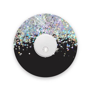 May include: A round, black object with a white, circular component in the center. The top half is covered in silver glitter, with a gradient effect. The glitter has a holographic sheen, reflecting multiple colors.