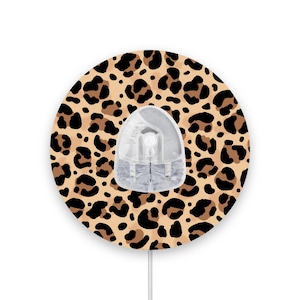 May include: A round, leopard print sticker for a medical device. The sticker is brown and black with a clear plastic cover.
