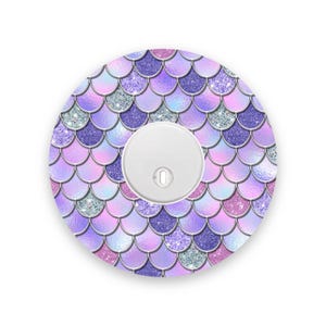 May include: A round, white object with a central button, surrounded by a mermaid scale design. The scales are in shades of purple, pink, and silver glitter, creating a shimmering effect. The design is on a white background.