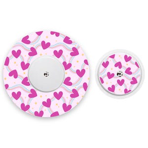 May include: Two medical device covers with a pink heart pattern and rainbow accents. The larger cover has a white center with a black design, and the smaller cover mirrors the design. The background is white.