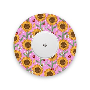 May include: A circular medical device with a white central disc, set against a pink backdrop adorned with sunflowers, bees, and ladybirds. The sunflowers have yellow petals and brown centres. The bees and ladybirds are also visible.