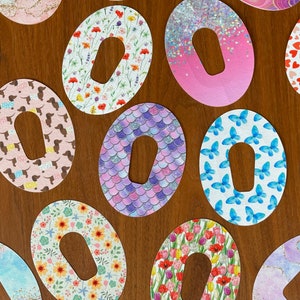 以下が含まれることがあります： 中央に穴が開いた、カラフルで模様のある粘着包帯の詰め合わせ。包帯には、蝶、花、ハート、キラキラなどのデザインが施されています。