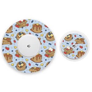 May include: Two round, adhesive, medical device covers with a blue background and a pattern of cartoon pancakes, waffles, strawberries, and blueberries. The larger cover has a central hole for a medical device.