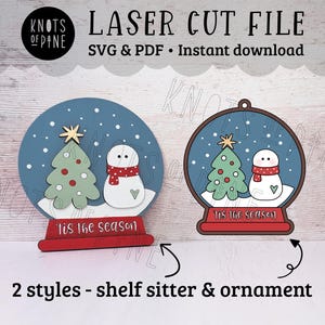 May include: Laser cut file for a snow globe ornament with a snowman and a Christmas tree inside. The snow globe is blue with white snowflakes and the text "Tis the season" on the base. The file is available in SVG and PDF formats.