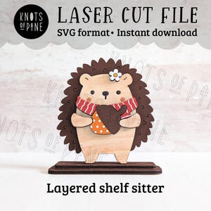 May include: A layered shelf sitter featuring a wooden hedgehog with a brown spiky back, wearing a scarf and holding an acorn. The design includes the text "LASER CUT FILE" and "SVG format • Instant download."