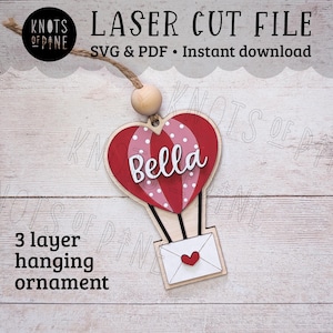 Può includere: Un ornamento appeso in legno a tre strati tagliato al laser a forma di mongolfiera con un cuore rosso e pois bianchi. Il pallone ha la scritta "Bella" e una piccola busta con un cuore appesa in basso.
