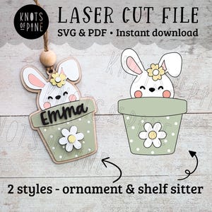May include: Two styles of laser cut bunny in a pot ornament. One is a hanging ornament and the other is a shelf sitter. Both have a green pot with white polka dots and a white flower with a yellow center. The bunny is white with pink cheeks and a yellow flower on its head. The hanging ornament has the name "Emma" written on the pot.