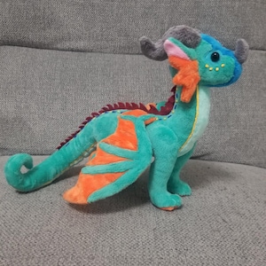 May include: A teal green plush dragon with orange wings and a gray horn. The dragon has yellow eyes and a pink mouth.