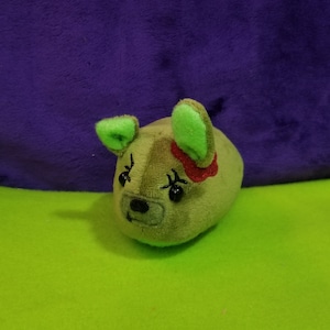 May include: A plush toy in the shape of a small animal, with a rounded body and green ears. The toy is olive green with a black nose and eyes, and a red accent. It is set against a green and purple background.