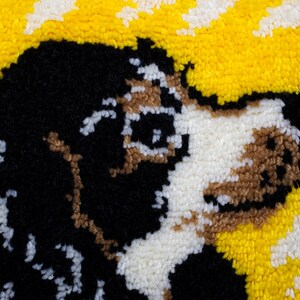 May include: A close-up of a handmade rug depicting a dog's face. The dog has black, white, and brown fur, set against a yellow and white background. The rug is made with a textured, looped pile.