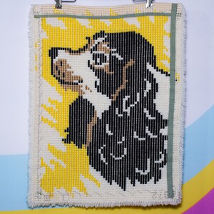 May include: A handmade tapestry depicting a dog's profile. The dog is rendered in black, white, and tan yarn against a yellow backdrop. The rectangular tapestry has a cream border and a green ribbon along the top edge.