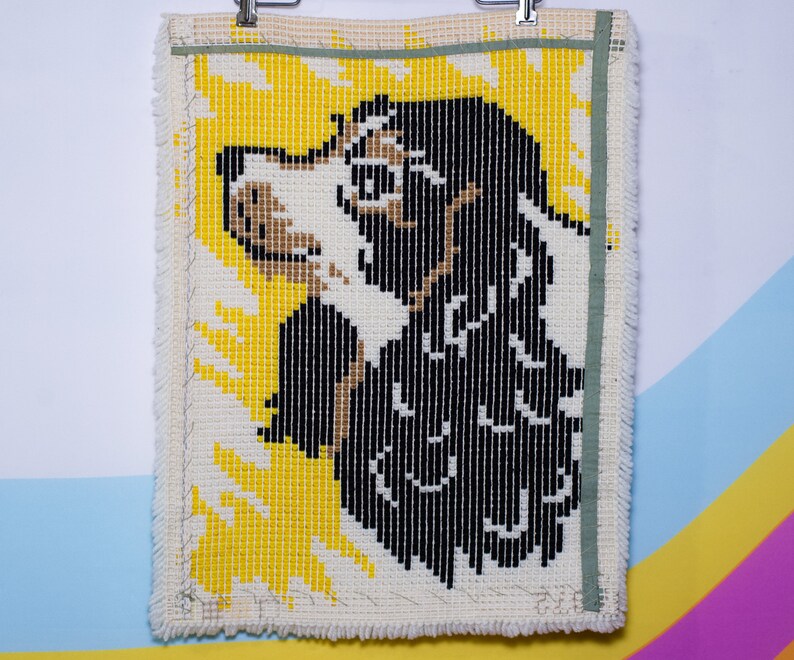 May include: A handmade tapestry depicting a dog's profile. The dog is rendered in black, white, and tan yarn against a yellow backdrop. The rectangular tapestry has a cream border and a green ribbon along the top edge.