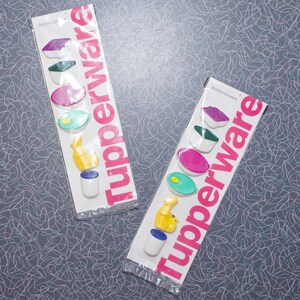 May include: Two packages of Tupperware button covers. Each package contains six colorful button covers in various shapes and colors, including purple, green, pink, and yellow. The Tupperware logo is printed in pink on the packaging.