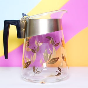 Peut inclure: Une carafe en verre transparent avec un bord et une poignée dorés. La carafe présente un motif de feuilles dorées et une poignée noire. Idéale pour servir des boissons, elle évoque un style vintage.