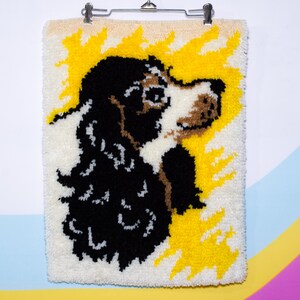 May include: A textured wall hanging with a dog's profile. The dog is rendered in black, brown, and white against a white backdrop, with yellow bursts of colour. The artwork is clipped to a metal hanger.