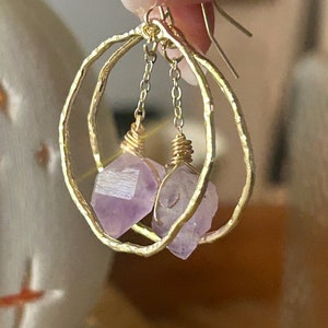 May include: A pair of gold-toned earrings featuring a hammered, circular hoop frame. Suspended from the center of each hoop by a short gold-toned chain is a raw, faceted amethyst crystal point, secured with gold-toned wire wrapping at the top.