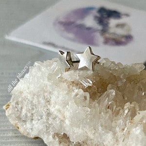 May include: A pair of silver star-shaped earrings on a white crystal background. The earrings are small and delicate, and they have a simple, elegant design.