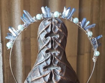 May include: A silver wire halo with blue crystal points and white beads. The halo is worn on a wooden statue.