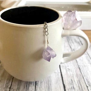 May include: A white ceramic mug with a silver chain and a purple amethyst crystal attached to it. The crystal is hanging from the chain inside the mug.