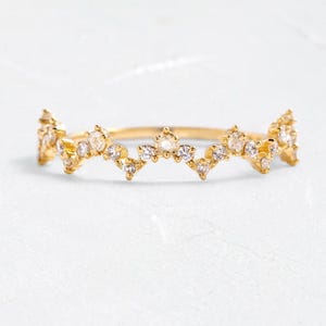Può includere: Un delicato anello d'oro con un design a forma di corona. L'anello presenta una serie di piccole gemme trasparenti sulla parte superiore, creando un effetto scintillante. La fascia è sottile e lucida, e l'anello è su uno sfondo bianco.