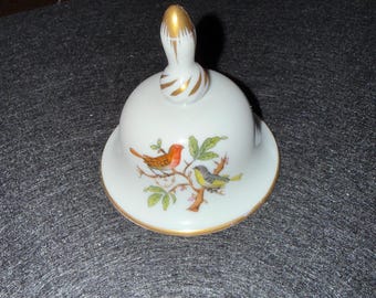 May include: A white ceramic bell with a gold-painted handle and rim. The bell features a painted design of two birds perched on a branch with green leaves. The birds are orange and yellow.