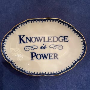May include: A white oval dish with a blue and gold trim. The dish has the words "Knowledge is Power" written on it in blue.
