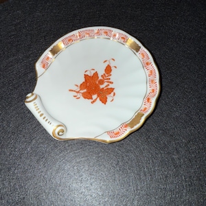 May include: A white porcelain shell-shaped dish with orange floral designs and gold trim. The dish has a rolled handle and a scalloped edge. The floral design is in the center of the dish.