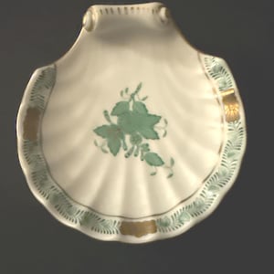 May include: A white, shell-shaped dish with a scalloped edge. The dish features a green floral design in the center and a green and gold border. The dish has a gold trim around the edge.