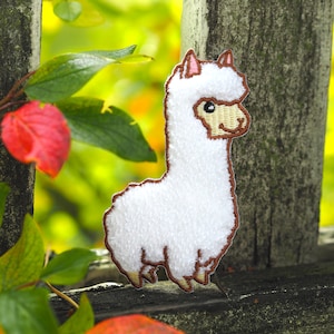Op de afbeelding: Witte geborduurde alpaca-applicatie met bruine stiksels. De alpaca staat op een houten oppervlak met groen gebladerte op de achtergrond.