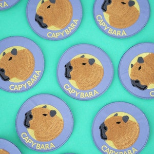 Może przedstawiać: Haftowane naszywki z motywem kapibary. Każda naszywka ma jasnofioletową obwódkę ze słowem "CAPYBARA" w kolorze żółtym. Kapibara jest brązowa z czarnymi detalami nosa i uszu. Naszywki są okrągłe.