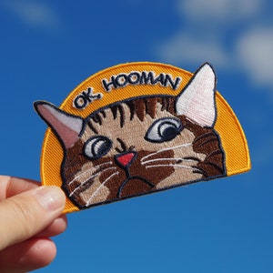 Op de afbeelding: Een geborduurde patch in geel en bruin met een kattenkop en de tekst "OK HOOMAN".