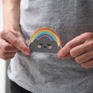 May include: A grey cloud with a rainbow over it and a smiling face. The cloud has closed eyes and pink cheeks.