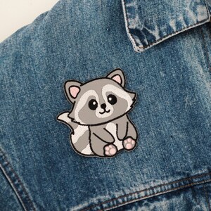 Könnte beinhalten: Ein gestickter Waschbär-Patch in Grau und Weiß mit rosa Akzenten. Der Patch ist auf eine blaue Jeansjacke genäht.