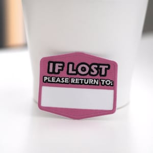 Può includere: Una toppa ricamata rosa e bianca con la scritta "IF LOST PLEASE RETURN TO:" in nero. La toppa ha uno spazio rettangolare bianco per la personalizzazione.