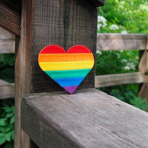 Könnte beinhalten: Ein regenbogenfarbener herzförmiger Patch ist an einem Holzlattenzaun befestigt. Der Patch ist aus Stoff und hat eine gewebte Textur.