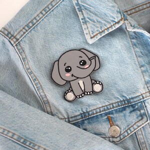 Könnte beinhalten: Ein gestickter grauer Elefanten-Patch mit weißen Akzenten und großen Augen. Der Patch ist auf eine blaue Jeansjacke gebügelt.