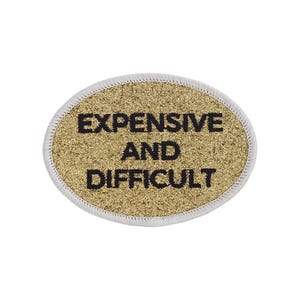 Op de afbeelding: Goudkleurige ovale patch met witte stiksels rond de randen. De patch heeft zwarte geborduurde tekst die "EXPENSIVE AND DIFFICULT" luidt.