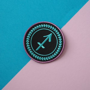 Op de afbeelding: Een geborduurde zwarte en turquoise patch met het sterrenbeeld Boogschutter in een roze en witte krans.