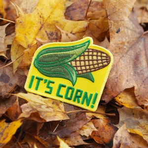 Pode incluir: Um remendo bordado amarelo com uma espiga de milho verde e marrom e o texto "IT'S CORN!" em um leito de folhas de outono.