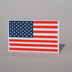 Könnte beinhalten: Ein kleiner, rechteckiger amerikanischer Flaggen-Patch mit roten und weißen Streifen und einem blauen Sternenfeld. Der Patch ist aus Stoff und hat einen weißen Rand.