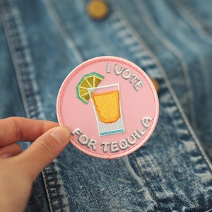 Może przedstawiać: Różowa, okrągła naszywka z haftem z tekstem "I vote for tequila" i kreskówkowym obrazkiem kieliszka do shotów z plasterkiem limonki.