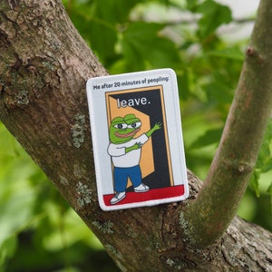 Op de afbeelding: Een witte patch met een groene cartoon kikker die een wit shirt en een blauwe spijkerbroek draagt. De kikker staat in een deuropening met het woord "leave." erboven geschreven. De tekst "Me after 20 minutes of peopling:" staat bovenaan de patch.