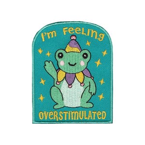 Puede incluir: Parche bordado con una rana verde con sombrero y cuello de bufón, con el texto "I'm Feeling Overstimulated". El parche tiene un fondo turquesa con estrellas amarillas.