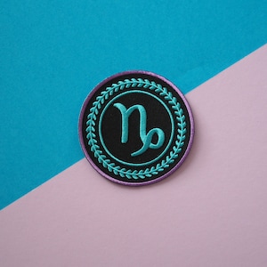 Op de afbeelding: Een geborduurde patch in turquoise en paars met een zwarte achtergrond met het astrologische symbool voor Steenbok.