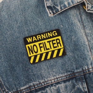 Könnte beinhalten: Ein gelber und schwarzer Warnschild-Patch, der auf eine blaue Jeansjacke genäht ist. Das Schild lautet "WARNING NO FILTER".